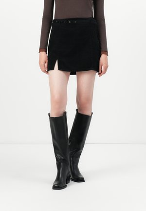 Svart kordfløyels miniskjørt med en frontsplit, kombinert med høye svarte lærstøvler med bred legg og chunky hæl.