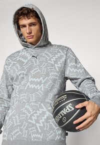 Ung mand i en grå mønstret hættetrøje, der holder en sort Spalding basketball mod en lys ensfarvet baggrund.