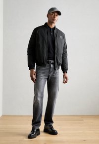 Svart bomberjakke, svart poloskjorte, grå jeans, svarte snørestøvler og en grå caps. Uformell antrekk på tre-gulv mot en lys bakgrunn.