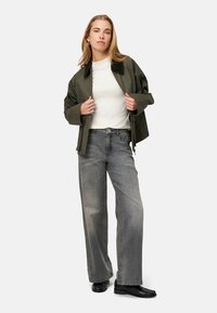 Olivově zelená bunda s černým límcem, pod ní bílá košile a šedé džíny s širokými nohavicemi. Outfit doplňují černé mokasíny.