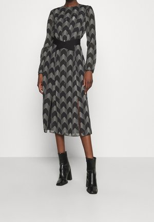 Femme portant une robe midi à manches longues avec un motif chevron noir et blanc, des fentes latérales et des bottines noires à talons.