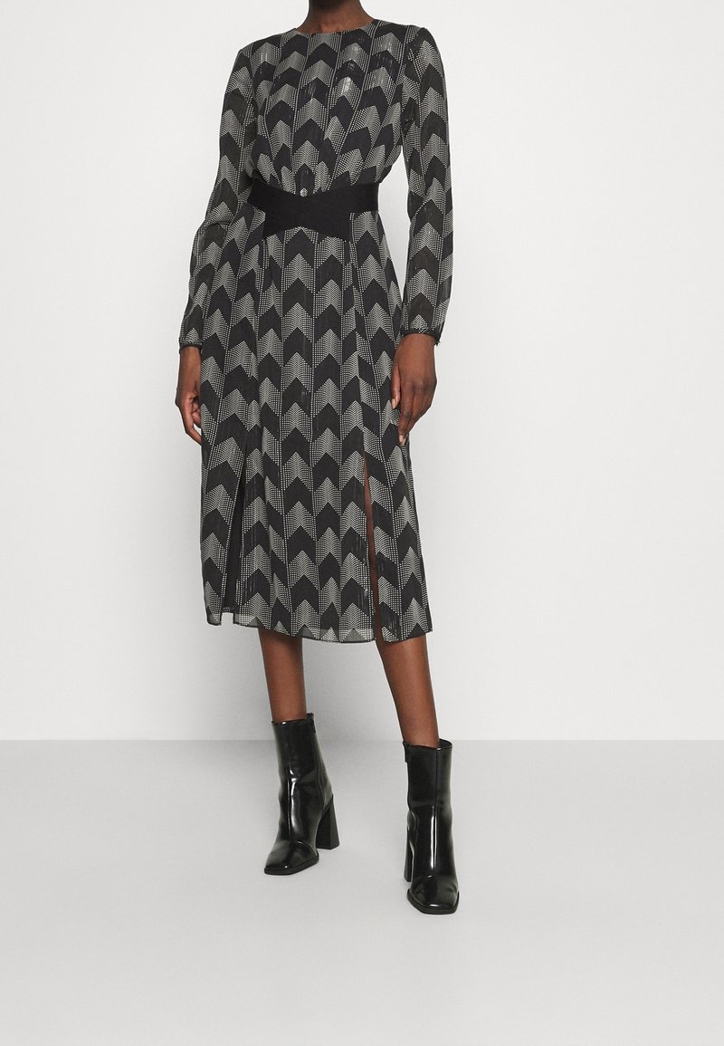 Femme portant une robe midi à manches longues avec un motif chevron noir et blanc, des fentes latérales et des bottines noires à talons.
