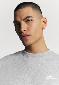 Felpa grigia con scollo a girocollo e tessuto testurizzato. Presenta un piccolo logo Nike bianco sul petto. Capelli corti, orecchino visibile.