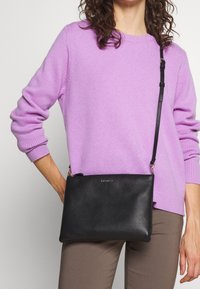 Černá kožená kabelka přes rameno s texturovaným povrchem, zlatými doplňky a nastavitelním popruhem, kterou drží osoba oblečená v levandulovém svetru.