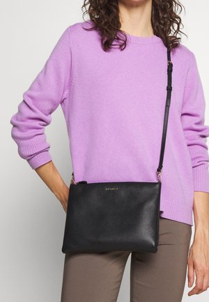 Černá kožená kabelka přes rameno s texturovaným povrchem, zlatými doplňky a nastavitelním popruhem, kterou drží osoba oblečená v levandulovém svetru.