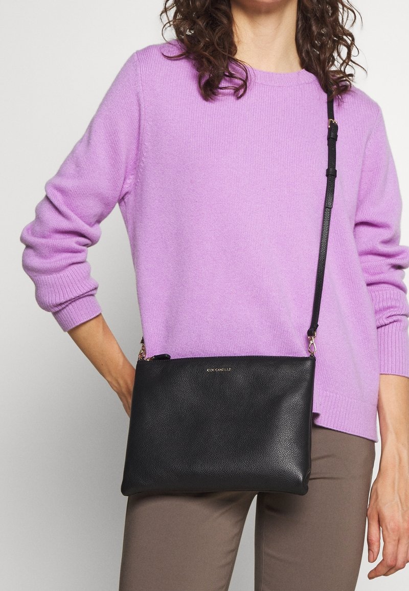 Černá kožená kabelka přes rameno s texturovaným povrchem, zlatými doplňky a nastavitelním popruhem, kterou drží osoba oblečená v levandulovém svetru.