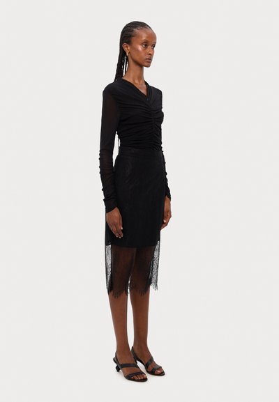 Blusa negra de manga larga con detalle fruncido en la parte frontal, combinada con una falda negra con encaje. La modelo lleva sandalias negras de tiras.