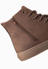 Zapatilla alta marrón hecha de cuero sintético, con una cremallera lateral, cordones redondos y una suela de goma. Presenta una textura suave y costuras reforzadas.