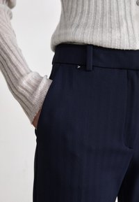 Pantaloni blu navy con motivo a spina di pesce, dotati di un logo sottile sulla vita e una texture liscia. Abbinati a un maglione leggero a coste.