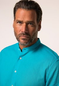 Camicia color turchese con chiusura a bottoni, realizzata in un tessuto morbido; presenta un colletto alto e una vestibilità rilassata.