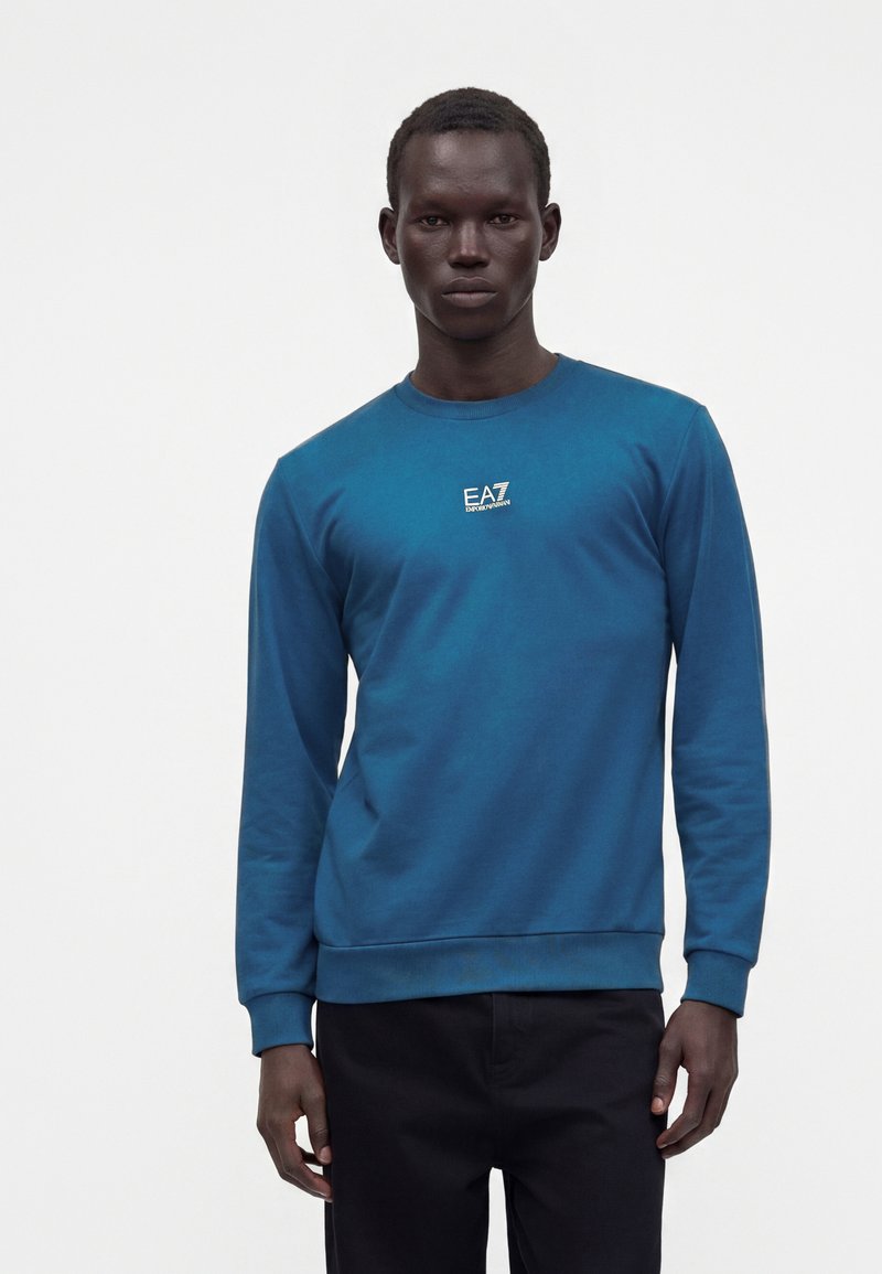 Homme portant un sweat-shirt bleu à manches longues avec le logo blanc "EA7 Emporio Armani" et un pantalon noir, debout devant un fond blanc uni.