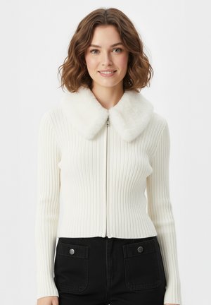 Maglione bianco a coste con zip, caratterizzato da un morbido colletto in pelliccia sintetica e maniche lunghe, indossato con pantaloni neri decorati con bottoni.