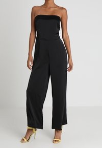 Combinaison noire sans bretelles avec un corsage ajusté et un pantalon à jambe large, fabriquée en tissu lisse. Associée à des sandales à talons hauts jaunes.