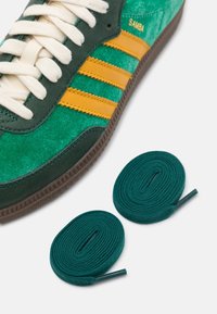 Baskets en suède vert avec des rayures jaunes, lacets crème et semelle en caoutchouc marron. Deux lacets teal assortis placés à côté des baskets.