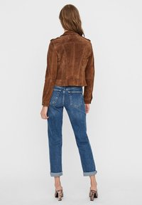 Korte bruine suede jas met puntige kraag en schouderepauletten, gedragen met hoge taille blauwe jeans en slangenpatroon hakken.