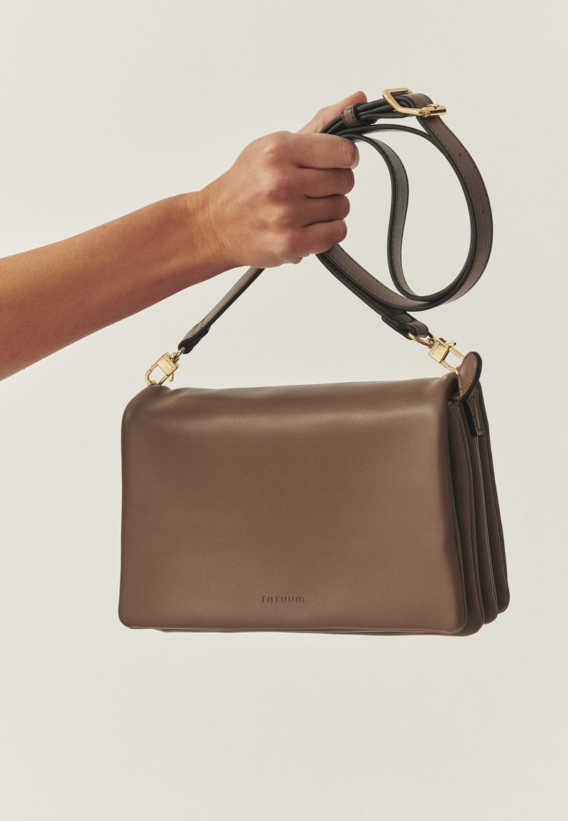 Kéz barna bőr válltáskával, aranyszínű fémalkatrészekkel és állítható pánttal, egyszínű bézs háttér előtt.