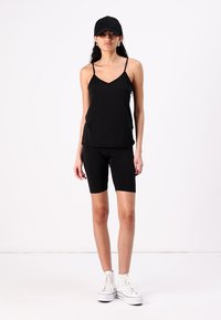 Top preto sem mangas com alças finas, combinado com calções de bicicleta pretos e ténis brancos de cano alto. Um boné preto completa o visual.