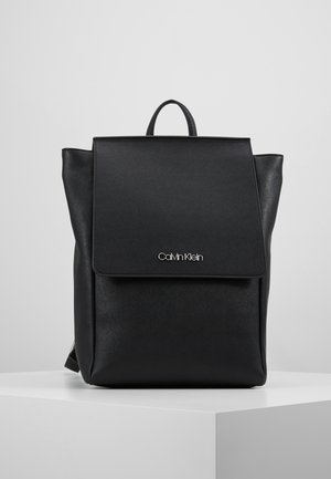 Sac à dos noir Calvin Klein avec rabat avant et poignée supérieure, présenté sur un piédestal blanc sur un fond clair uni.