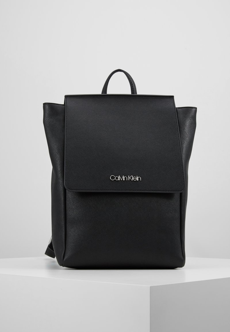 Sac à dos noir Calvin Klein avec rabat avant et poignée supérieure, présenté sur un piédestal blanc sur un fond clair uni.