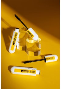 Deux tubes de mascara avec des caps en relief blancs et des étiquettes jaunes, l'un étiqueté "Bounce" et l'autre "theCOLLOSAL (CURL)", sur un fond jaune.
