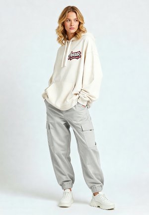 Felpa oversize beige con grafica "Amour", abbinata a pantaloni cargo grigi chiari e sneakers bianche. Tessuto morbido, vestibilità rilassata, tasche visibili.