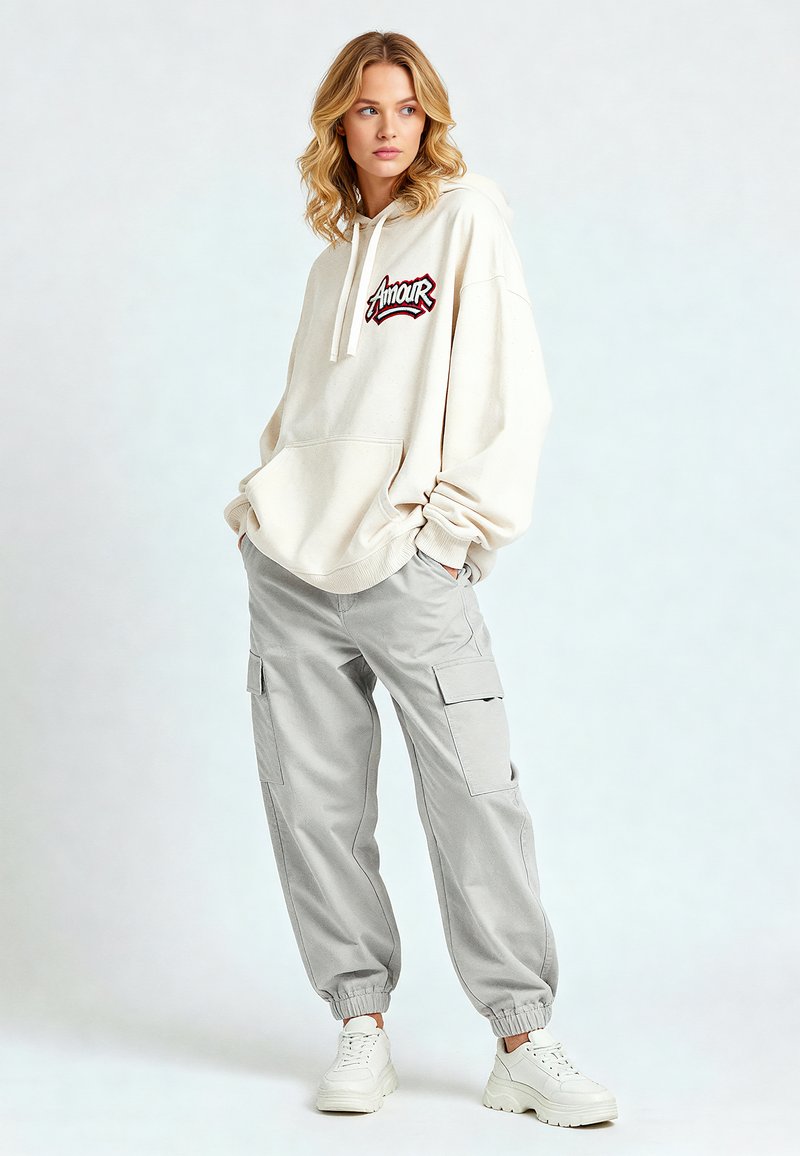 Felpa oversize beige con grafica "Amour", abbinata a pantaloni cargo grigi chiari e sneakers bianche. Tessuto morbido, vestibilità rilassata, tasche visibili.