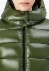 Pufferjack in olijfgroen met een hoge kraag, glanzende textuur en zwarte rits. Heeft drukknoopjes langs de kraag.