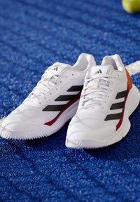 Witte sportschoenen met zwarte en rode accenten, een bovenwerk van mesh, een gestructureerde zool en het drie strepen-logo. Geplaatst op een blauwe, gestructureerde ondergrond.