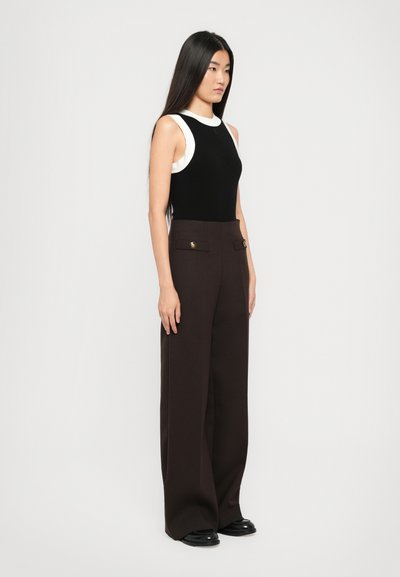 Vrouw met lang zwart haar, gekleed in een mouwloze zwart-witte top, hoge smoezelige donkerbruine wijde broek en zwarte schoenen, staande in zijaanzicht.