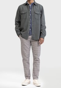 Grijze shirtjack met twee borstzakken over een blauwe bloes. Lichtgrijze geblokte broek, witte sneakers. Gladde stoftextuur.