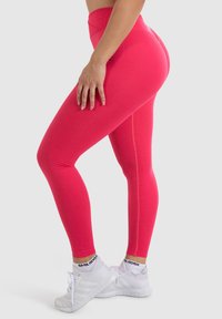 Leggings aderenti a vita alta di un brillante colore rosa, realizzati in materiale elastico. Presentano una texture liscia e una vita a coste, abbinati a sneakers bianche.