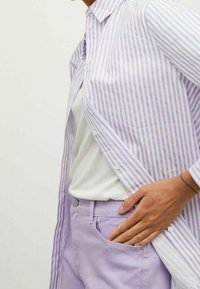 Personne portant une chemise rayée violet clair sur un haut blanc avec un pantalon violet clair, la main reposant dans la poche du pantalon.
