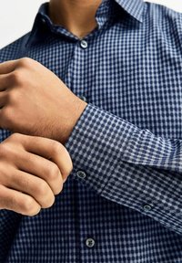 Niebiesko-biała kraciaste koszula na guziki z długimi rękawami, z mankietem, który jest regulowany, oraz jasnoniebieskimi guzikami. Gładka tekstura tkaniny.