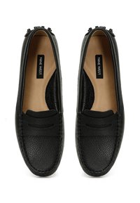 Mocasines de cuero negro con una textura granulada, que presentan detalles cosidos y un diseño sin cordones. El interior es beige y cuenta con una etiqueta de la marca.