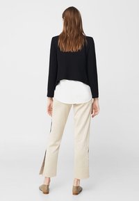 Veste courte noire portée sur une chemise blanche, associée à un pantalon beige avec des fentes sur les côtés, et des chaussures grises sans lacets. Design simple et minimaliste.