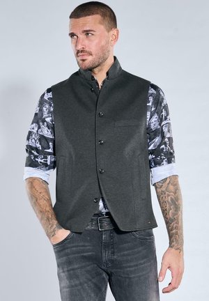 Dunkelgraue Weste mit fünf Knöpfen, schlanker Passform und einer Brusttasche, getragen über einem gemusterten schwarz-weißen Hemd, kombiniert mit grauen Jeans.