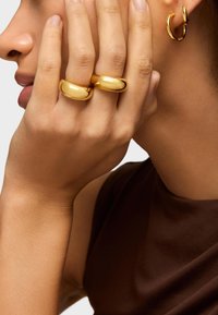 Dwa złote pierścionki i mały złoty kolczyk w kształcie kółka. Gładka, błyszcząca powierzchnia i zaokrąglone kształty. Ciepłe odcienie skóry w zestawieniu z ciemnym topem.