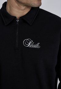 Polo negro con cremallera y cuello doblado. Presenta un logo bordado blanco en el pecho. La tela parece suave y texturizada.