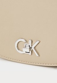 Borsa a tracolla in pelle beige con un clasps in argento raffigurante la scritta "CK". La superficie ha una finitura testurizzata e cuciture a contrasto.