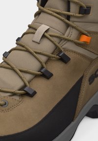 Botte de randonnée marron clair et noire avec languette rembourrée, lacets bruns passant par des œillets noirs et un œillet crochet orange.