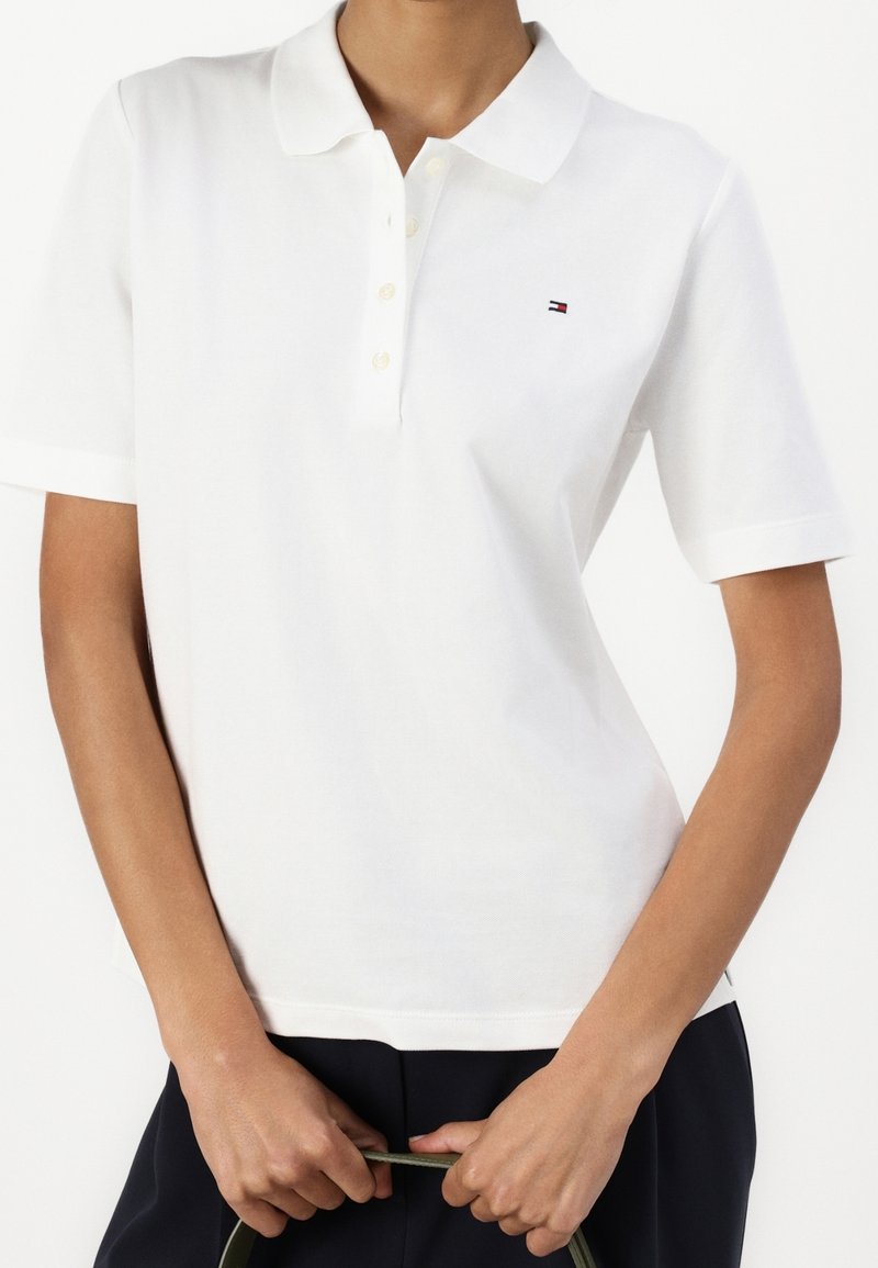 Personne portant un polo blanc à manches courtes avec un petit logo sur la poitrine, tenant une sangle ou une poignée avec les deux mains.