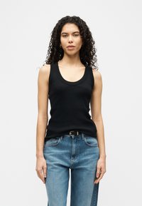 Femeie cu păr creț, purtând un top negru fără mâneci, tucked în blugi albaștri cu o curea neagră, stând pe un fundal alb simplu.