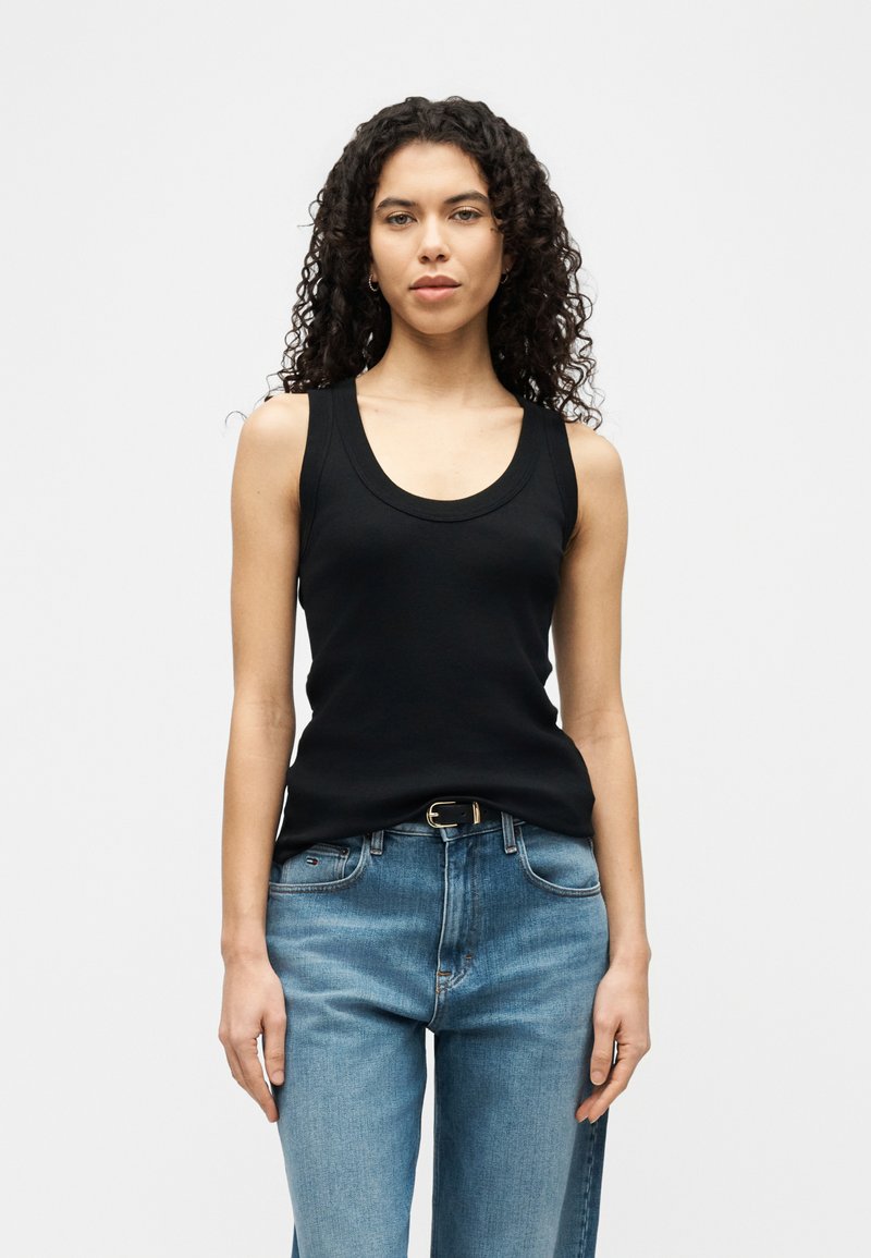 Femeie cu păr creț, purtând un top negru fără mâneci, tucked în blugi albaștri cu o curea neagră, stând pe un fundal alb simplu.