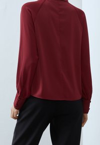Blusa borgogna a maniche lunghe realizzata in tessuto morbido, con polsini abbottonati e orlo dritto, vista da dietro su uno sfondo chiaro.
