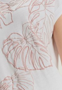 Biała tkanina z wypukłymi, różowymi haftowanymi konturami dużych tropikalnych liści na górnej części bezrękawnik.