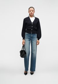 Woman with braided hair wearing black cardigan, white top, blue jeans, black pointed shoes, holding black handbag, standing against plain background.