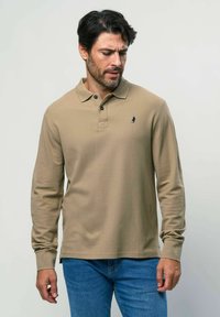 Beež pikkade varrukatega polo särk klassikalise kraega, kahe nupuga kinnitusega ja tumesinise logoga. Sile puuvillane kangas, koos siniste teksadega.