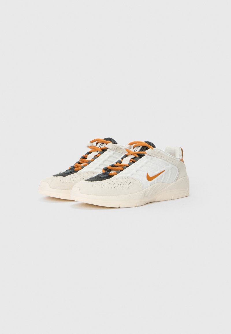 Para białych i beżowych sneakersów z pomarańczowym logo Nike i pomarańczowymi sznurówkami, na jednolitym jasnym tle.
