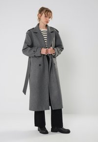 Cappotto grigio in misto lana con design doppiopetto, ampi revers e cintura. Presenta grandi bottoni e dettagli sulle spalle. Abbinato a un top a righe.