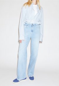 Person iført lyseblå brede ben jeans, blå slip-on sko og en hvid t-shirt med en blå og hvid stribet skjorte.
